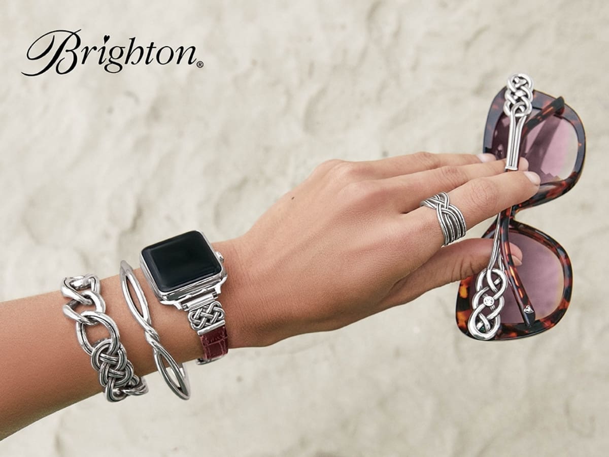 A hand adorned with silver jewelry and a smartwatch, holding a pair of sunglasses against a textured background with the word "brighton.