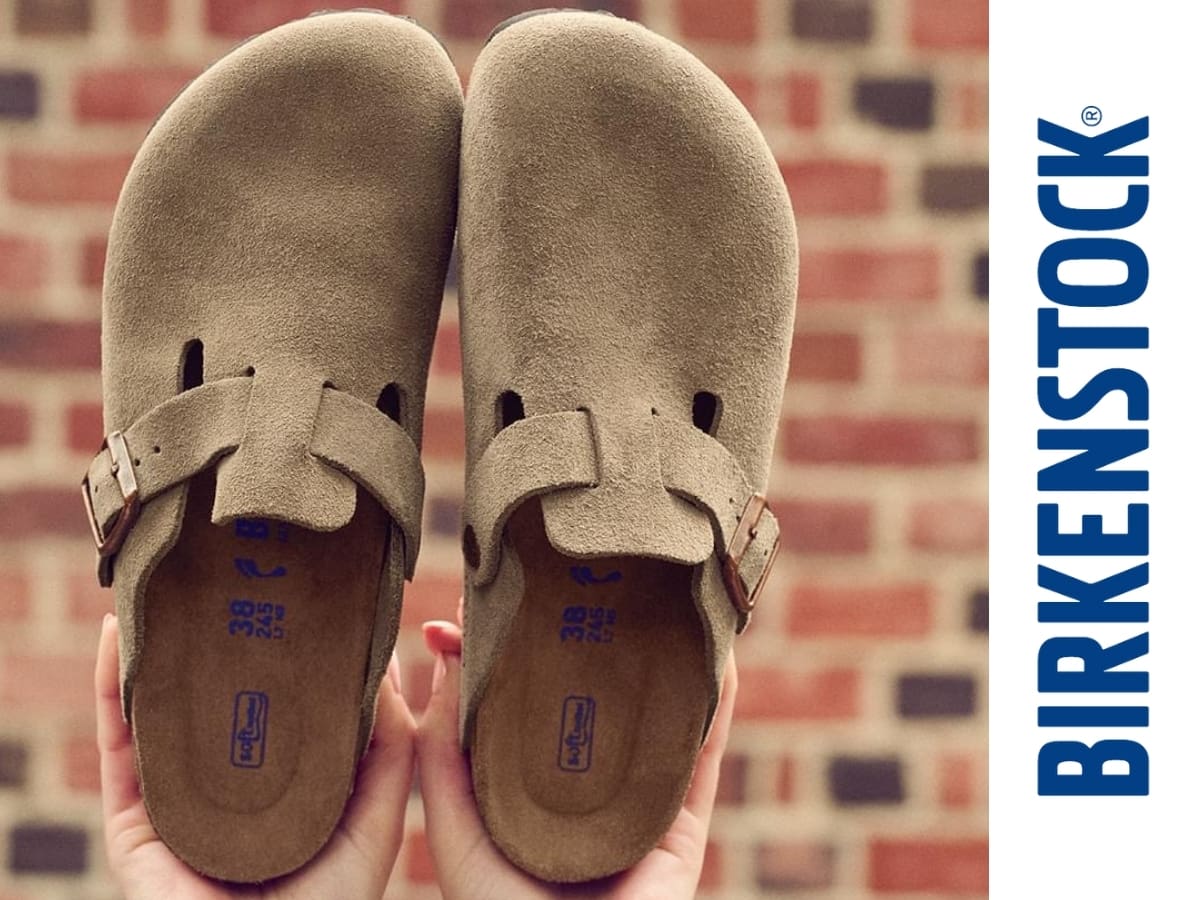 A pair of birkenstock sandals being held up in front of a brick wall.