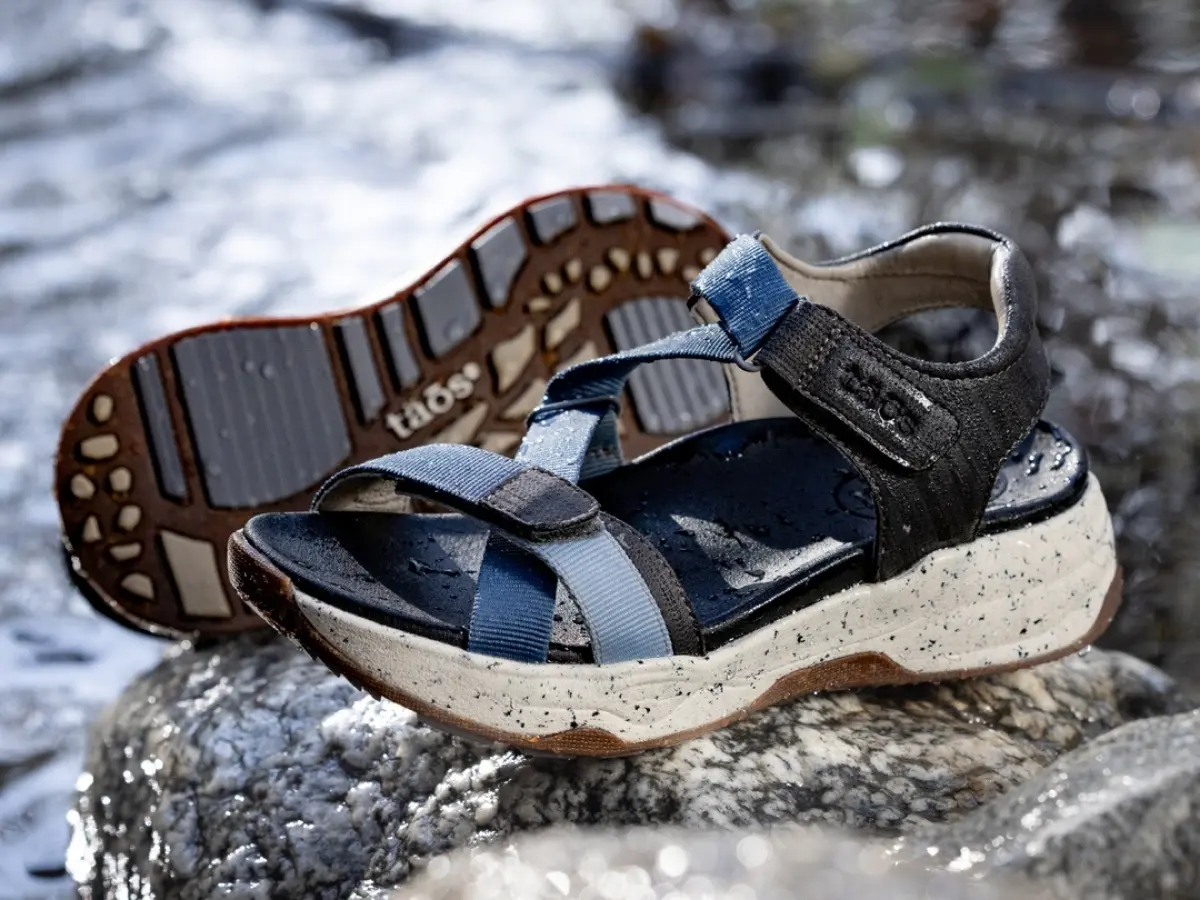 A pair of outdoor sandals placed on a rock near water, highlighting their rugged soles.